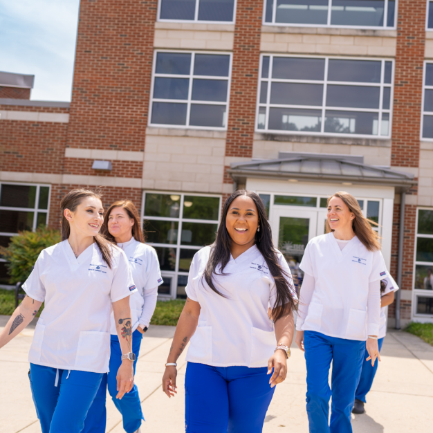 A group of women in scrubs walking on a sidewalk outside of a campus building.