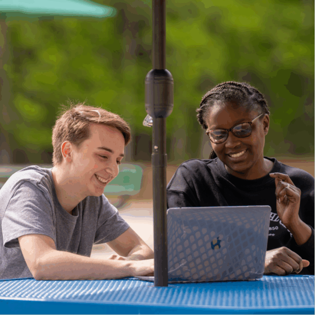 Two people seated at an outdoor table under an umbrella using a laptop.