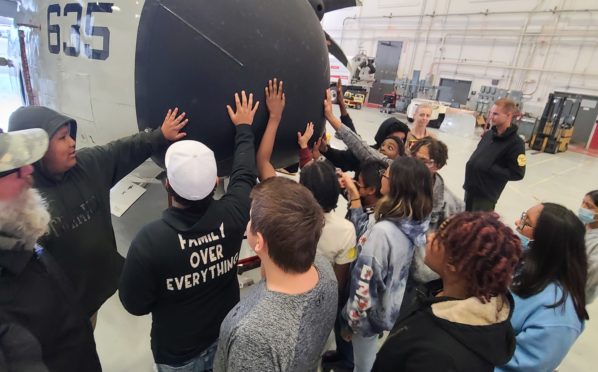 A group of people gathered around the nose of an aircraft in a hanger.