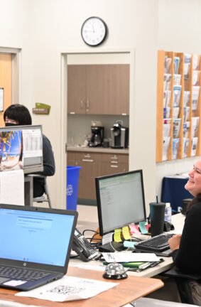 A woman standing in front of a desk with another woman seated behind it using a computer