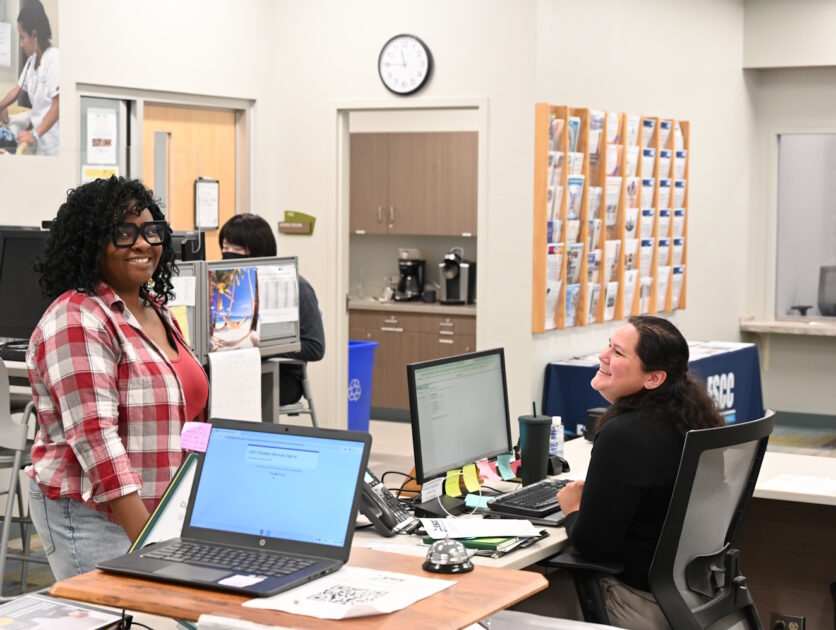 A woman standing in front of a desk with another woman seated behind it using a computer