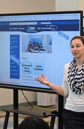 A woman standing beside a large monitor giving a presentation