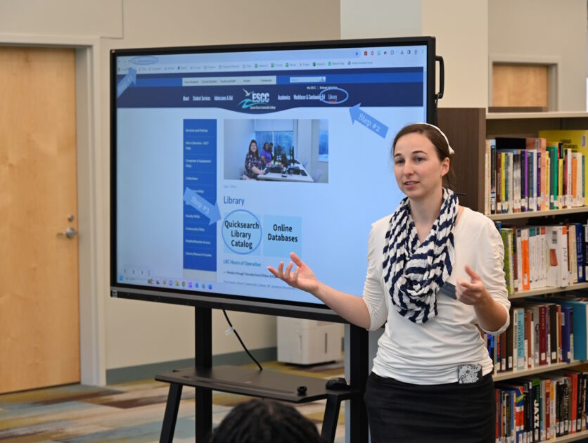 A woman standing beside a large monitor giving a presentation