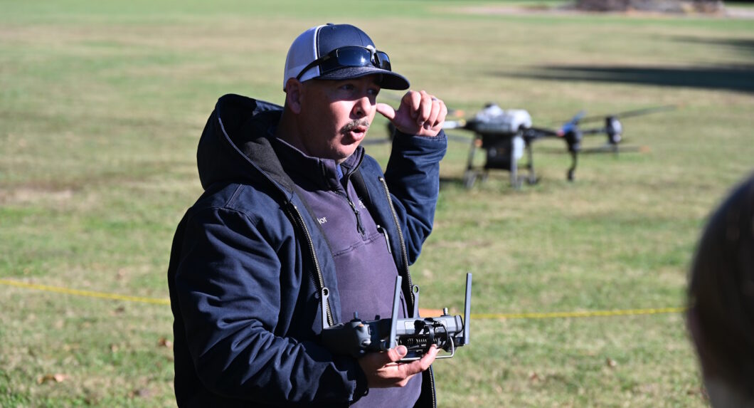 A man holding a drone remote, wearing a ball cap, and pointing back over his shoulder