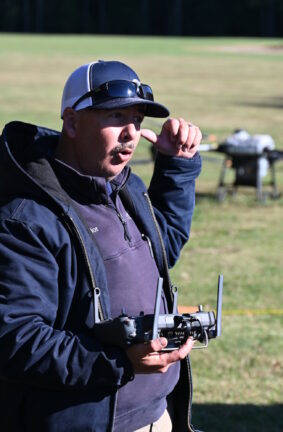 A man holding a drone remote, wearing a ball cap, and pointing back over his shoulder
