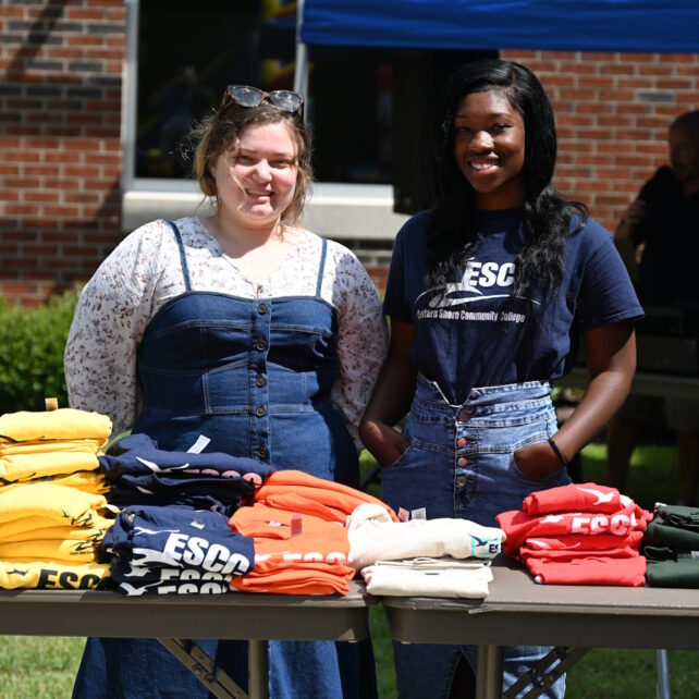 Two women standing outside behind a table with stacks of t-shirts folded on it.