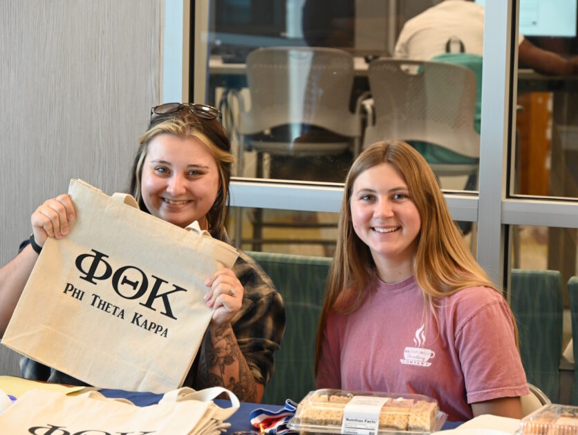 Two women seated behind a table. One is holding up a bag with Phi Theta Kappa on it.