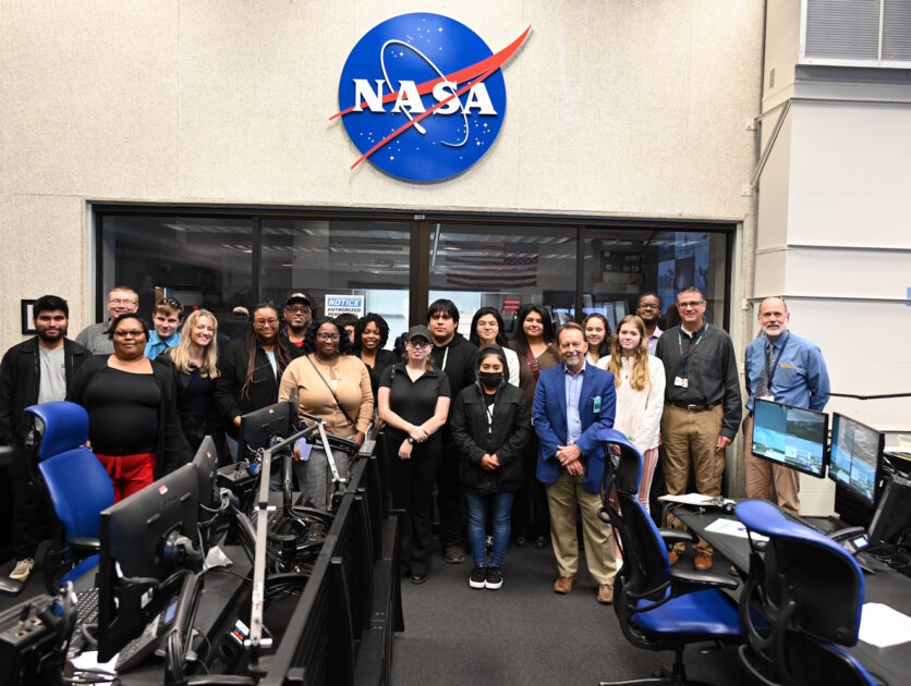 A group of people standing in front of rows of computers under a NASA sign.