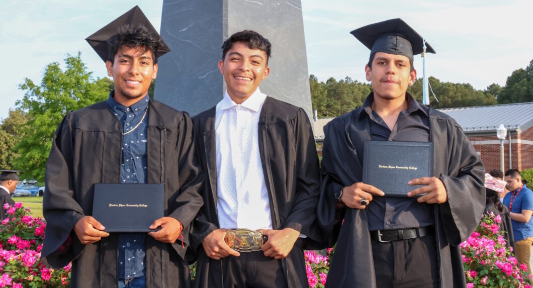 Three men wearing graduation caps and gowns, holding their diplomas and standing outside.