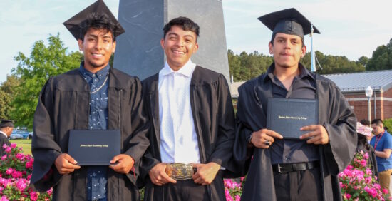 Three men wearing graduation caps and gowns, holding their diplomas and standing outside.