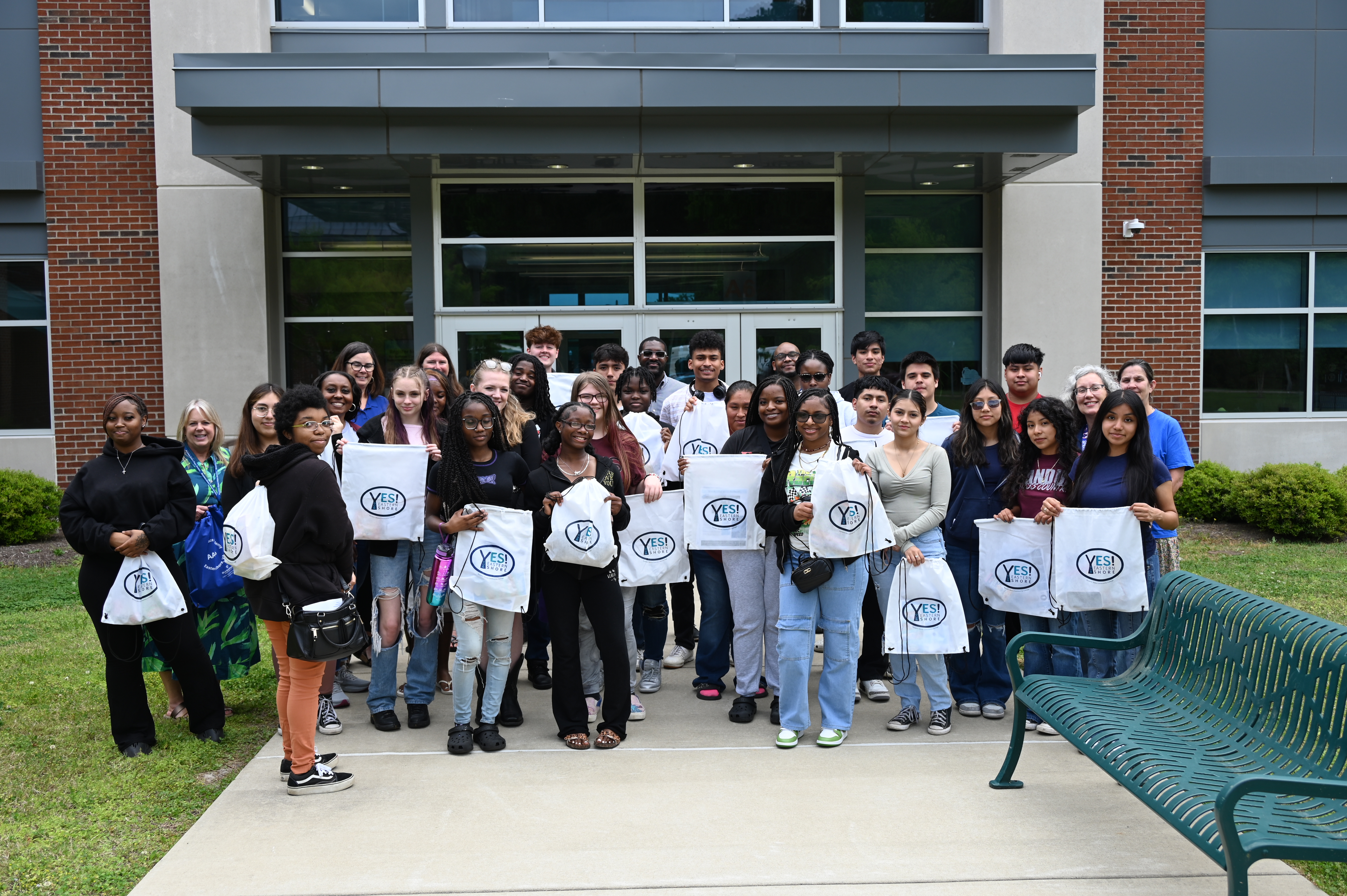A large group do people standing outside in front of a campus building holding plastic bags saying YES Eastern Shore on them.