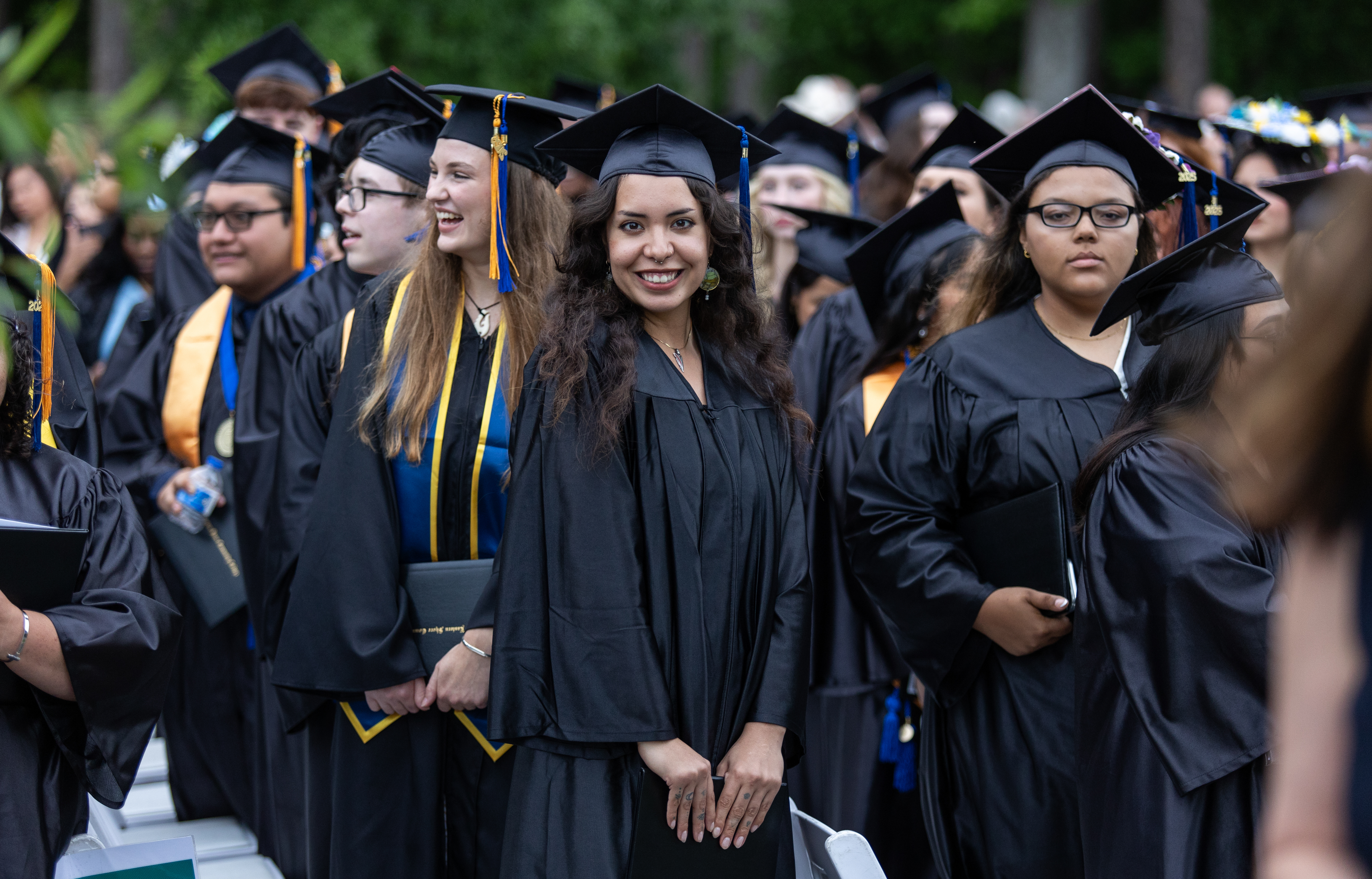 A close-up shot of graduating students standing outside in their black academic gowns and caps. In the center, a female student with long dark hair and gold honor cords smiles brightly at the camera, flanked by other graduates lined up for the ceremony.