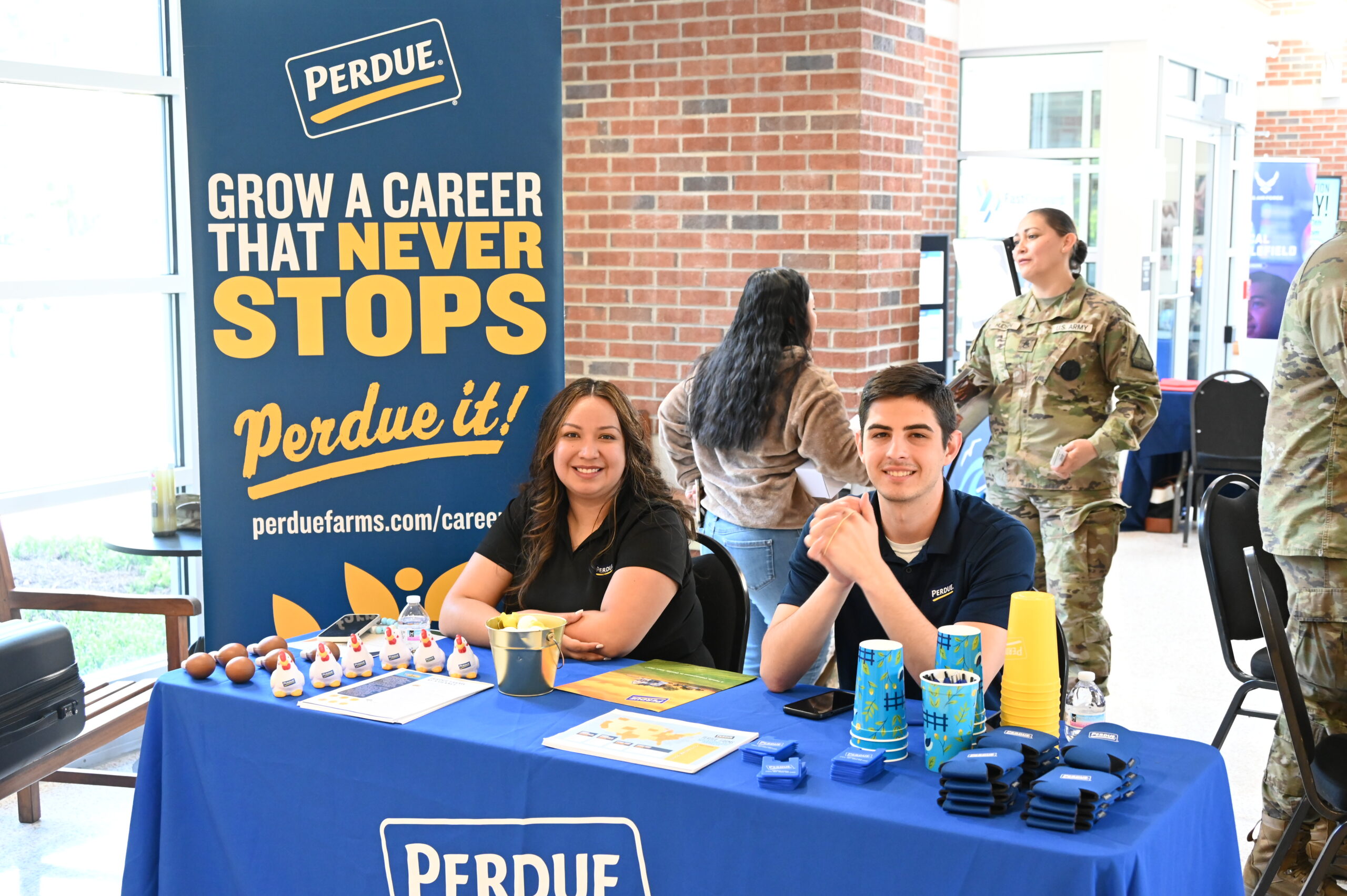 A young man and woman smile while sitting behind a blue table at a Perdue Farms career booth indoors. Next to them is a tall banner that reads, "GROW A CAREER THAT NEVER STOPS. Perdue it! perduefarms.com/careers". In the background, other event attendees and an individual in a military uniform are visible.
