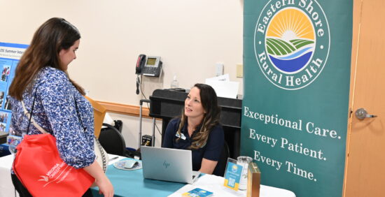 A young woman with a red Virginia Career Works tote bag over her shoulder stands at an event table, speaking with a representative. Behind the table is a large banner for Eastern Shore Rural Health featuring their logo and the slogan, "Exceptional Care. Every Patient. Every Time