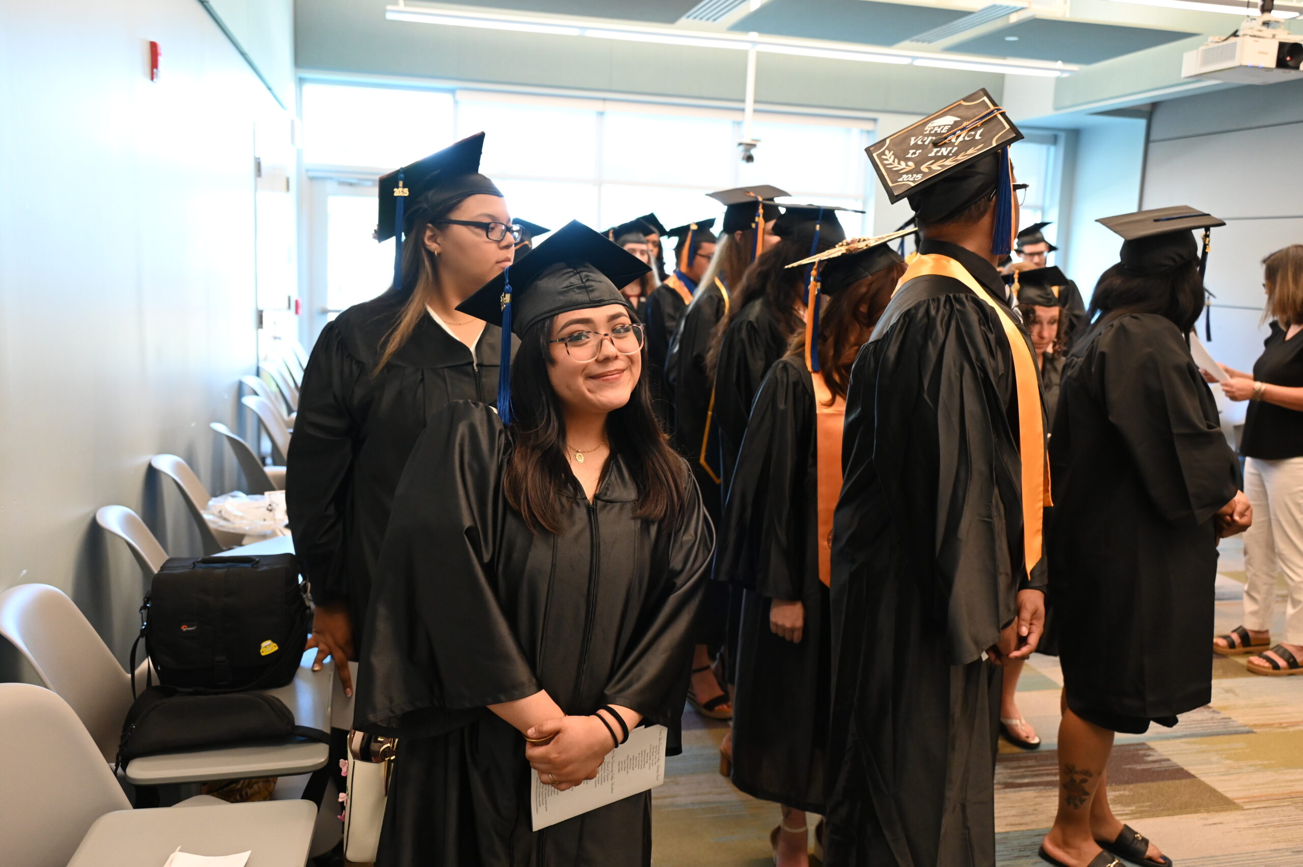 A group of graduating students dressed in black academic gowns and caps standing in a line indoors. A female student near the front smiles warmly toward the camera.