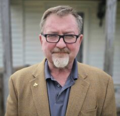 A middle-aged man with short brown hair and black rimmed glasses, wearing a tan blazer over a grey collared shirt.