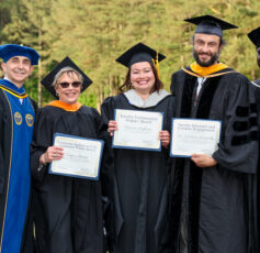 Five faculty members in graduation stand outdoors together at an academic ceremony, smiling and holding award certificates in front of a wooded background