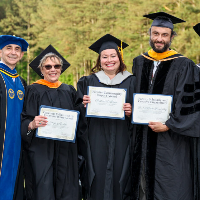 Five faculty members in graduation stand outdoors together at an academic ceremony, smiling and holding award certificates in front of a wooded background