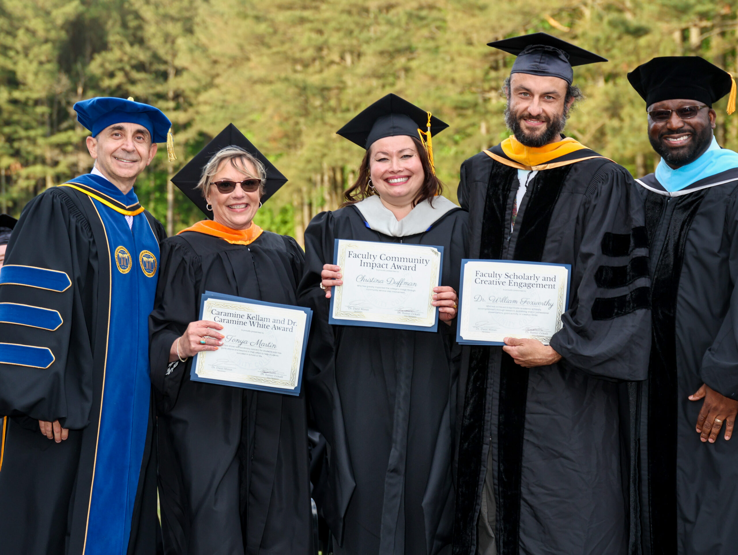 Five faculty members in graduation stand outdoors together at an academic ceremony, smiling and holding award certificates in front of a wooded background