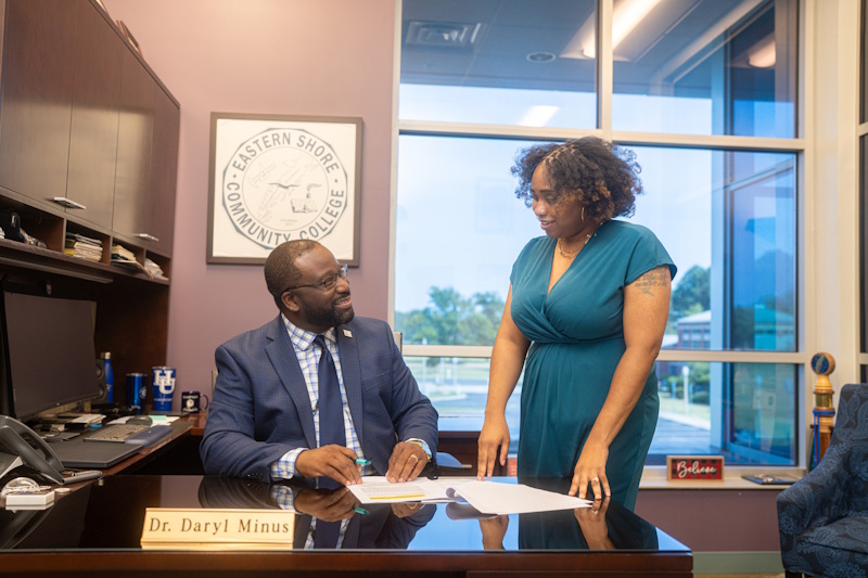 Dr. Daryl Minus seated at his desk speaking to a woman standing next to him.